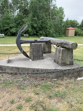 A large metal anchor and a thick wooden log are displayed on concrete pedestals, surrounded by a chain barrier. The scene is set in a grassy park area with lush green trees in the background. A small brick structure with a red roof stands nearby. The overall atmosphere is one of a historical or maritime display.