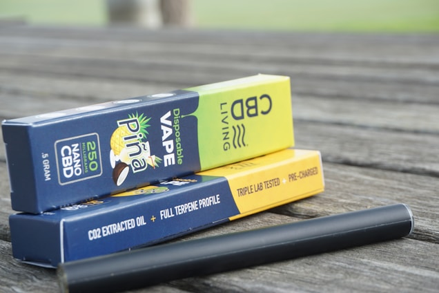 Boxes of disposable CBD vape products are stacked on a wooden surface. The packaging includes branding, flavor descriptions, and product details like '250 NANO CBD' and 'Disposable Vape'. A black vape pen is lying next to the boxes.