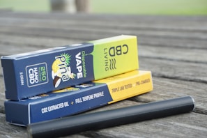 Boxes of disposable CBD vape products are stacked on a wooden surface. The packaging includes branding, flavor descriptions, and product details like '250 NANO CBD' and 'Disposable Vape'. A black vape pen is lying next to the boxes.