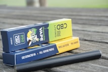 Boxes of disposable CBD vape products are stacked on a wooden surface. The packaging includes branding, flavor descriptions, and product details like '250 NANO CBD' and 'Disposable Vape'. A black vape pen is lying next to the boxes.