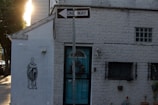 A brick building with a one-way sign above the entrance. On the left side of the building, there is a mural of a person in a robe holding an object. The building has barred windows and some small potted plants near the entrance. Sunlight is visible in the background, casting shadows on the façade.