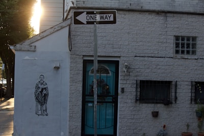 A brick building with a one-way sign above the entrance. On the left side of the building, there is a mural of a person in a robe holding an object. The building has barred windows and some small potted plants near the entrance. Sunlight is visible in the background, casting shadows on the façade.