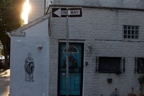 A brick building with a one-way sign above the entrance. On the left side of the building, there is a mural of a person in a robe holding an object. The building has barred windows and some small potted plants near the entrance. Sunlight is visible in the background, casting shadows on the façade.