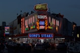 A brightly lit diner offers a lively scene with neon lights highlighting the name of the establishment, emphasizing steak offerings. The atmosphere is bustling with people gathered around despite the evening setting, and cars are parked in front of the establishment.
