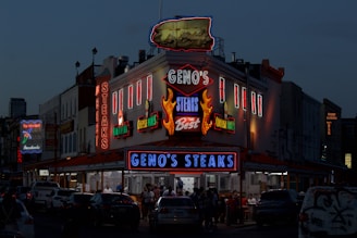 A brightly lit diner offers a lively scene with neon lights highlighting the name of the establishment, emphasizing steak offerings. The atmosphere is bustling with people gathered around despite the evening setting, and cars are parked in front of the establishment.