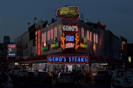 A brightly lit diner offers a lively scene with neon lights highlighting the name of the establishment, emphasizing steak offerings. The atmosphere is bustling with people gathered around despite the evening setting, and cars are parked in front of the establishment.