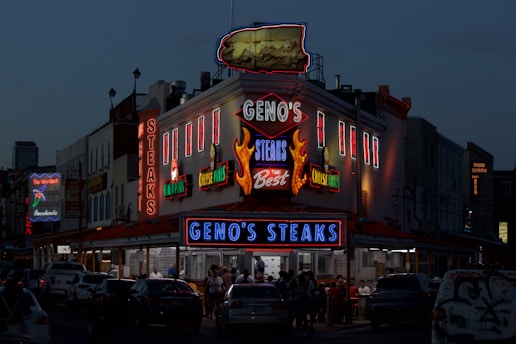A brightly lit diner offers a lively scene with neon lights highlighting the name of the establishment, emphasizing steak offerings. The atmosphere is bustling with people gathered around despite the evening setting, and cars are parked in front of the establishment.