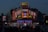 A brightly lit diner offers a lively scene with neon lights highlighting the name of the establishment, emphasizing steak offerings. The atmosphere is bustling with people gathered around despite the evening setting, and cars are parked in front of the establishment.