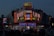 A brightly lit diner offers a lively scene with neon lights highlighting the name of the establishment, emphasizing steak offerings. The atmosphere is bustling with people gathered around despite the evening setting, and cars are parked in front of the establishment.