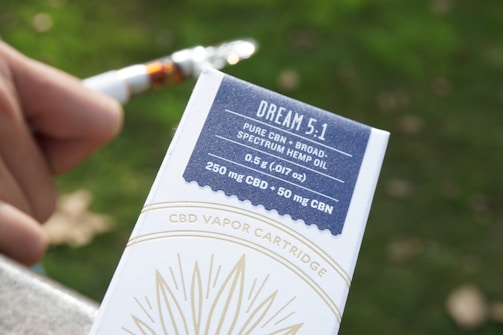 A close-up of a person's hand holding a CBD vapor cartridge package labeled 'DREAM 5.1' with details about the product including its contents (pure CBN, broad-spectrum hemp oil, 0.5 g, 250 mg CBD, 50 mg CBN) highlighted on a blue and white box. The background features a blurred green and brown outdoor environment.