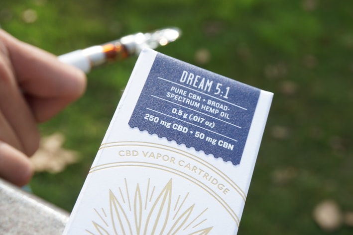 A close-up of a person's hand holding a CBD vapor cartridge package labeled 'DREAM 5.1' with details about the product including its contents (pure CBN, broad-spectrum hemp oil, 0.5 g, 250 mg CBD, 50 mg CBN) highlighted on a blue and white box. The background features a blurred green and brown outdoor environment.
