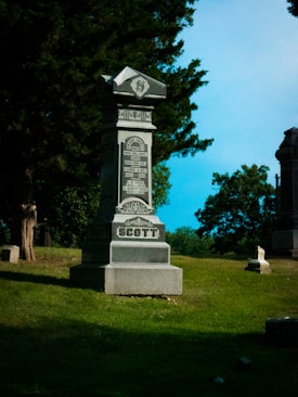 A large, ornate tombstone is situated in a sunlit cemetery, surrounded by lush green grass and trees. The name 'Scott' is engraved prominently on the base of the stone, with intricate details and inscriptions above. The sky is clear and blue, providing a serene backdrop.