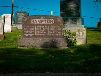 Multiple gravestones are situated in a cemetery, with inscriptions visible on a prominent stone in the foreground labeled 'HAMPTON'. The grass is well-maintained and the sky is blue, creating a serene setting.
