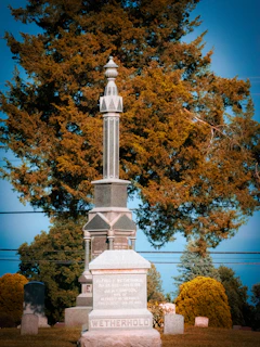A tall, ornate gravestone with intricate carvings stands prominently in a cemetery. The name 'Wetherhold' is inscribed on the base. In the background, large trees with autumn-colored leaves provide a natural backdrop, and a clear blue sky is visible.