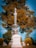 A tall, ornate gravestone with intricate carvings stands prominently in a cemetery. The name 'Wetherhold' is inscribed on the base. In the background, large trees with autumn-colored leaves provide a natural backdrop, and a clear blue sky is visible.