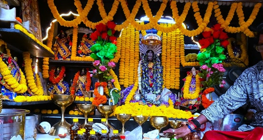 An elaborately decorated altar with statues adorned with vibrant garlands of yellow marigolds and colorful fabrics. Gold chalices are placed in front along with several bottles. Fresh flower arrangements with red and pink flowers add to the festive ambiance. The setting suggests a religious or ceremonial significance.