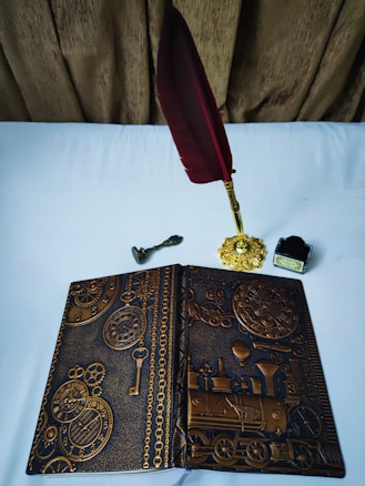 An ornately designed notebook with embossed gears, clocks, and a steam locomotive motif lies open on a white surface. Nearby, there is a vintage-style, gold-toned inkwell with a deep red feather quill and a small brass key.