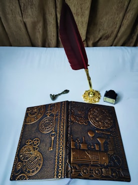 An ornately designed notebook with embossed gears, clocks, and a steam locomotive motif lies open on a white surface. Nearby, there is a vintage-style, gold-toned inkwell with a deep red feather quill and a small brass key.