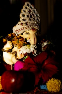 A detailed sculpture of a deity, adorned with an ornate headdress embellished with pearls and colorful beads. The statue is surrounded by vibrant flowers, including a prominent red hibiscus and other assorted blooms. The figure holds flowers in its hands, creating a harmonious blend of vivid colors and intricate craftsmanship.