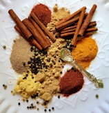 An assortment of spices including cinnamon sticks, ground spices, and various peppercorns is arranged on a white textured surface. A decorative golden spoon is also present among the spices.