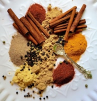 An assortment of spices including cinnamon sticks, ground spices, and various peppercorns is arranged on a white textured surface. A decorative golden spoon is also present among the spices.