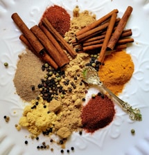 An assortment of spices including cinnamon sticks, ground spices, and various peppercorns is arranged on a white textured surface. A decorative golden spoon is also present among the spices.