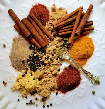 An assortment of spices including cinnamon sticks, ground spices, and various peppercorns is arranged on a white textured surface. A decorative golden spoon is also present among the spices.