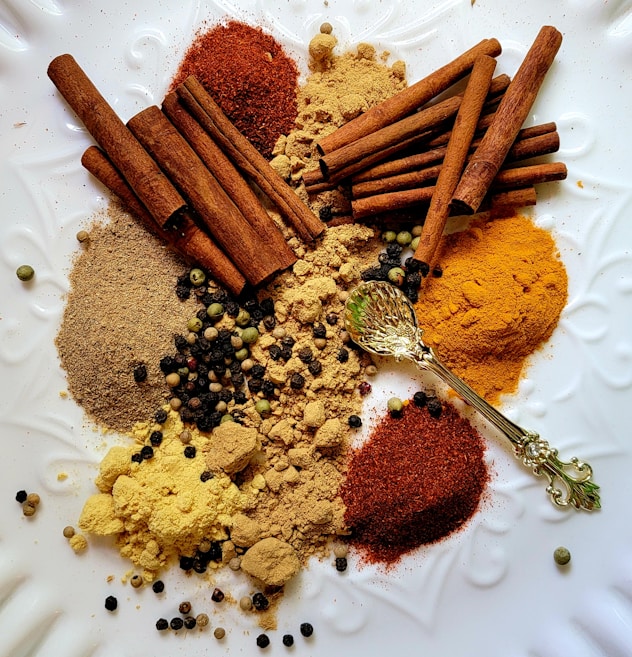 An assortment of spices including cinnamon sticks, ground spices, and various peppercorns is arranged on a white textured surface. A decorative golden spoon is also present among the spices.