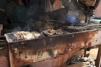An old, rusted barbecue grill with food cooking on it, emitting smoke. In the background, there are a few blurred objects and a person, but the focus remains on the grill and the food.