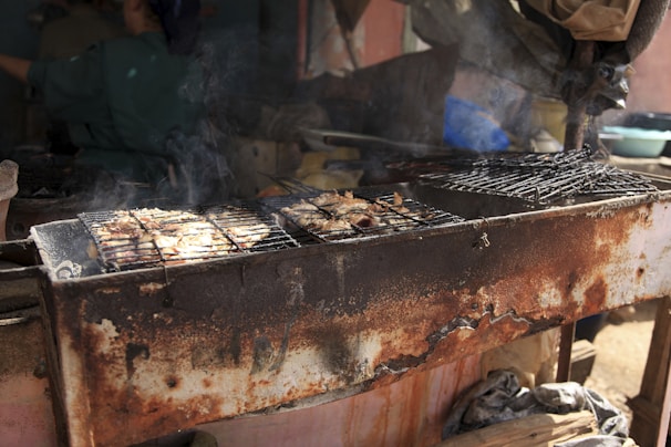 An old, rusted barbecue grill with food cooking on it, emitting smoke. In the background, there are a few blurred objects and a person, but the focus remains on the grill and the food.
