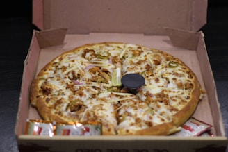 A freshly baked pizza with a golden crust is placed in an open cardboard box. It is topped with melted cheese, chunks of chicken, jalapeño slices, and some shaved onions. A small plastic table is in the center of the pizza, and condiment packets are placed beside it inside the box.