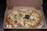 A freshly baked pizza with a golden crust is placed in an open cardboard box. It is topped with melted cheese, chunks of chicken, jalapeño slices, and some shaved onions. A small plastic table is in the center of the pizza, and condiment packets are placed beside it inside the box.