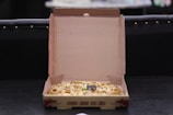 An open cardboard pizza box containing a pizza with visible toppings such as cheese and vegetables. The box is resting on a dark surface, and its interior is plain brown. The background is dimly lit, making the setting appear casual and relaxed.