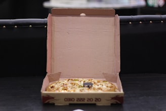 An open cardboard pizza box containing a pizza with visible toppings such as cheese and vegetables. The box is resting on a dark surface, and its interior is plain brown. The background is dimly lit, making the setting appear casual and relaxed.