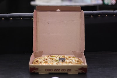 An open cardboard pizza box containing a pizza with visible toppings such as cheese and vegetables. The box is resting on a dark surface, and its interior is plain brown. The background is dimly lit, making the setting appear casual and relaxed.