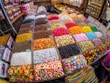 A vibrant market stall displaying an array of colorful candies and sweets in bins and trays. The sweets include various shapes and textures such as gummy bears, striped lollipops, and chocolate-coated treats, all neatly organized in individual sections.
