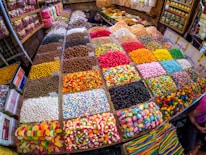 A vibrant market stall displaying an array of colorful candies and sweets in bins and trays. The sweets include various shapes and textures such as gummy bears, striped lollipops, and chocolate-coated treats, all neatly organized in individual sections.