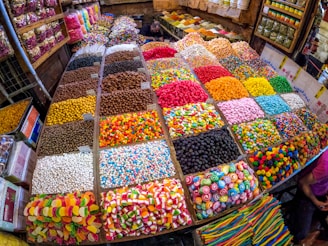 A vibrant market stall displaying an array of colorful candies and sweets in bins and trays. The sweets include various shapes and textures such as gummy bears, striped lollipops, and chocolate-coated treats, all neatly organized in individual sections.