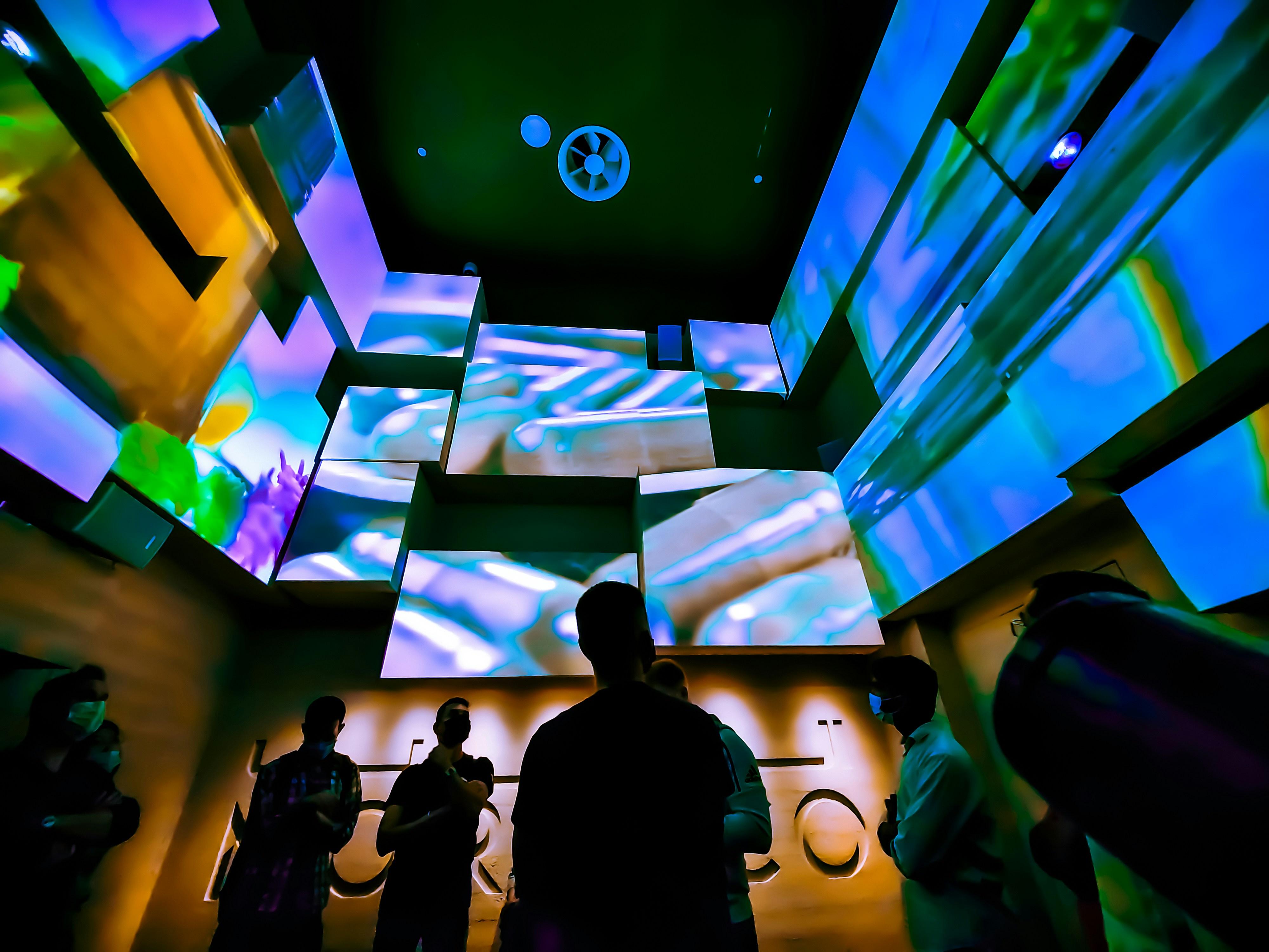 A group of people is gathered in a dimly lit room with colorful digital art projected on the walls. The walls are adorned with abstract patterns in vibrant blues, greens, and purples. The space feels modern and immersive, possibly resembling an art exhibition or a digital experience event.