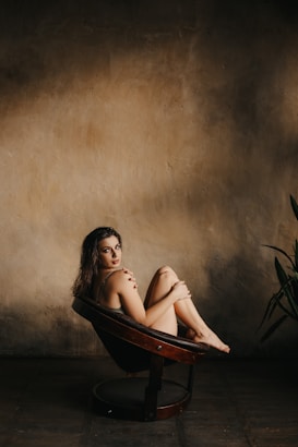 A woman is seated on a round, brown chair with her legs pulled close to her body. She is looking directly at the camera with a relaxed and contemplative expression. The background features a textured, warm-toned wall with soft lighting, casting gentle shadows and creating a serene atmosphere. Part of a plant is visible on the right side.