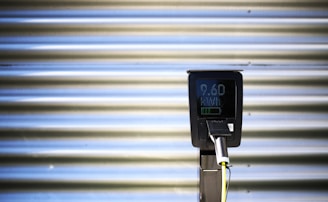A vertical black charging station with a digital screen is positioned against a corrugated metallic background. The screen displays numerical values, and there is a plugged-in charging cable hanging from the station.