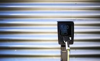 A vertical black charging station with a digital screen is positioned against a corrugated metallic background. The screen displays numerical values, and there is a plugged-in charging cable hanging from the station.