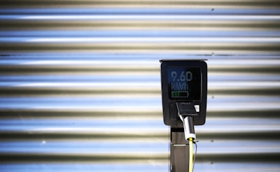 A vertical black charging station with a digital screen is positioned against a corrugated metallic background. The screen displays numerical values, and there is a plugged-in charging cable hanging from the station.
