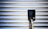 A vertical black charging station with a digital screen is positioned against a corrugated metallic background. The screen displays numerical values, and there is a plugged-in charging cable hanging from the station.