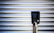 A vertical black charging station with a digital screen is positioned against a corrugated metallic background. The screen displays numerical values, and there is a plugged-in charging cable hanging from the station.