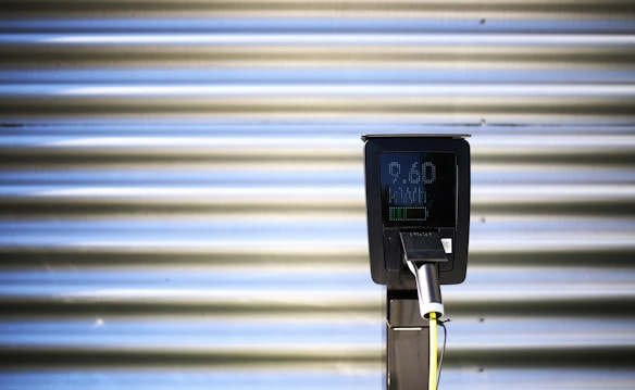 A vertical black charging station with a digital screen is positioned against a corrugated metallic background. The screen displays numerical values, and there is a plugged-in charging cable hanging from the station.