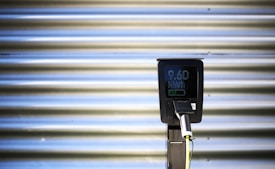 A vertical black charging station with a digital screen is positioned against a corrugated metallic background. The screen displays numerical values, and there is a plugged-in charging cable hanging from the station.