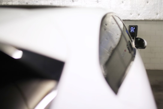 A close-up view of a white electric car parked in a garage, connected to a wall-mounted charging station that displays 25.6 kWh. The background consists of a textured, industrial-style wall.