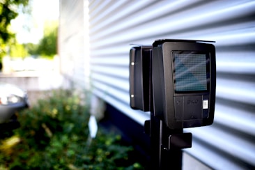 A close-up view of a black electronic device with a pixelated screen mounted on a stand in an outdoor environment. The background includes a corrugated metal wall and a blurry view of foliage and a parked car.