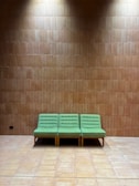 A row of three green upholstered chairs with button details sits against a brown tiled wall. The floor is also tiled, matching the wall color. The lighting casts a gentle shadow above the chairs, giving the setting a peaceful and minimalist feel.